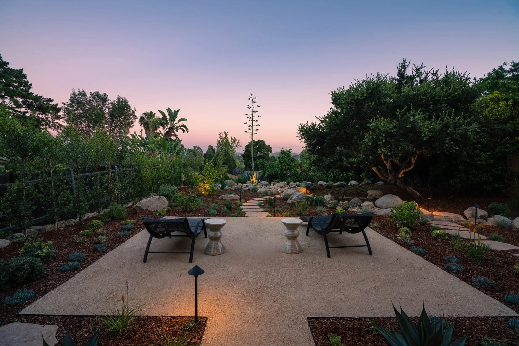 A gravel terrace with lounge chairs, stone paths, and illuminated planting at dusk.