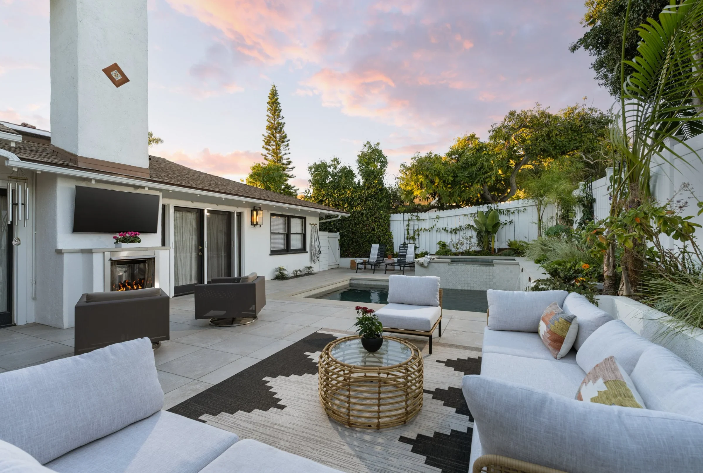 Pale seating, a woven table, and an outdoor fireplace beside a pool at sunset.