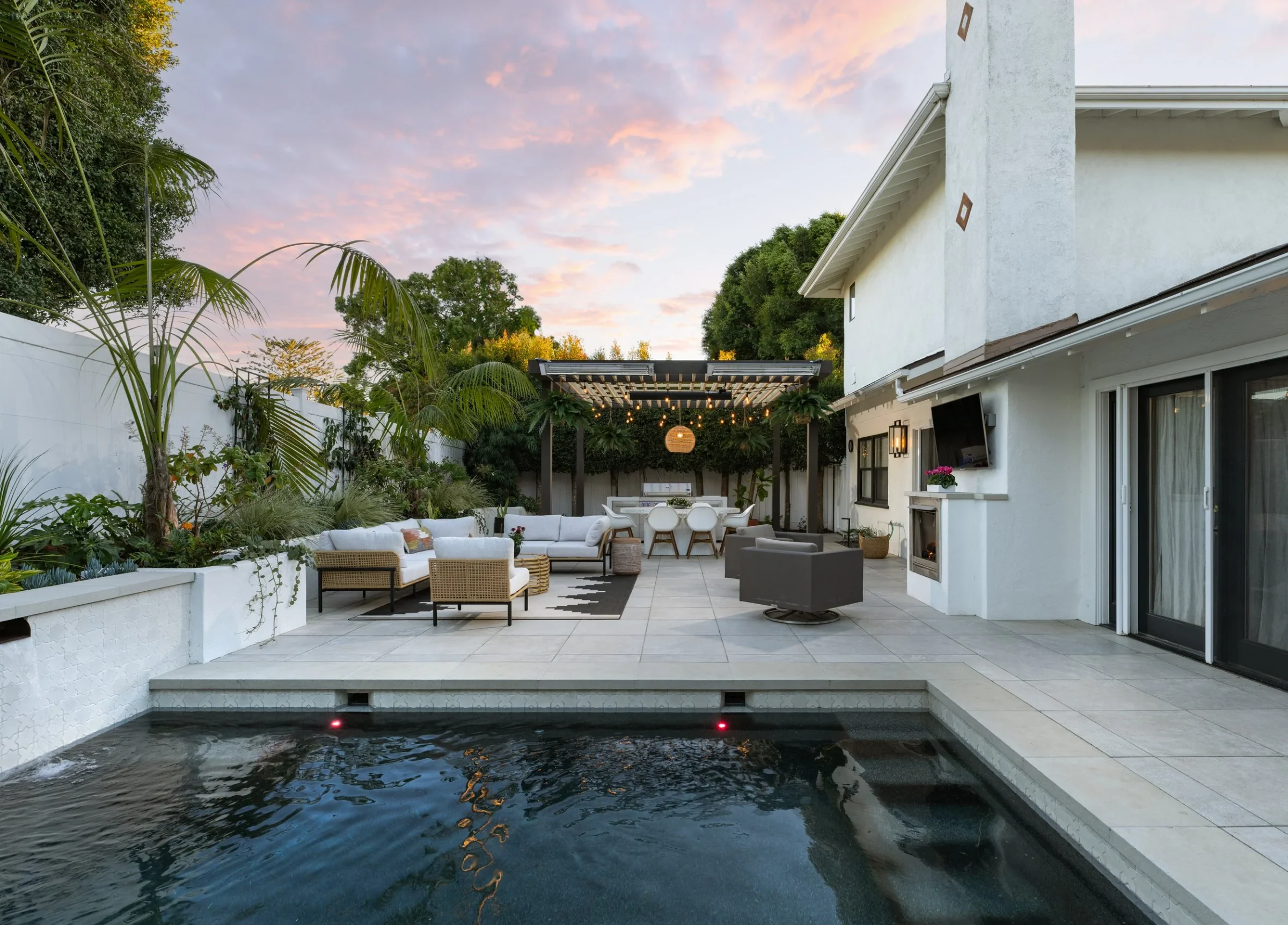 A view across a pool toward a pergola, planted borders, and an outdoor lounge.